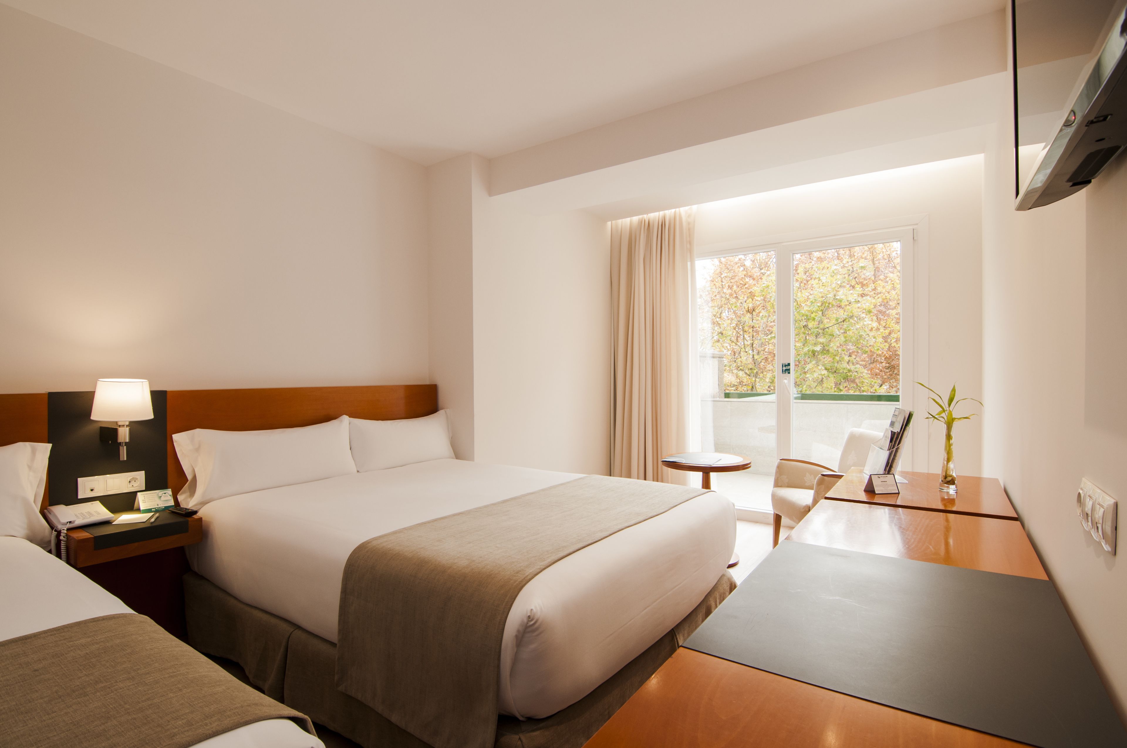 Habitación de hotel con una cama grande, mesita de noche con lámpara, una pequeña mesa redonda, una silla y un televisor en la pared.