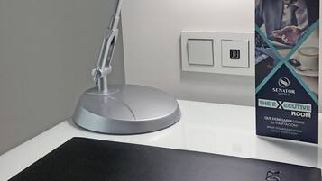 Une lampe de bureau moderne, dotée d’un fini métallique et d’un socle rectangulaire, est placée sur une surface blanche, à côté d’un interrupteur fixé au mur.