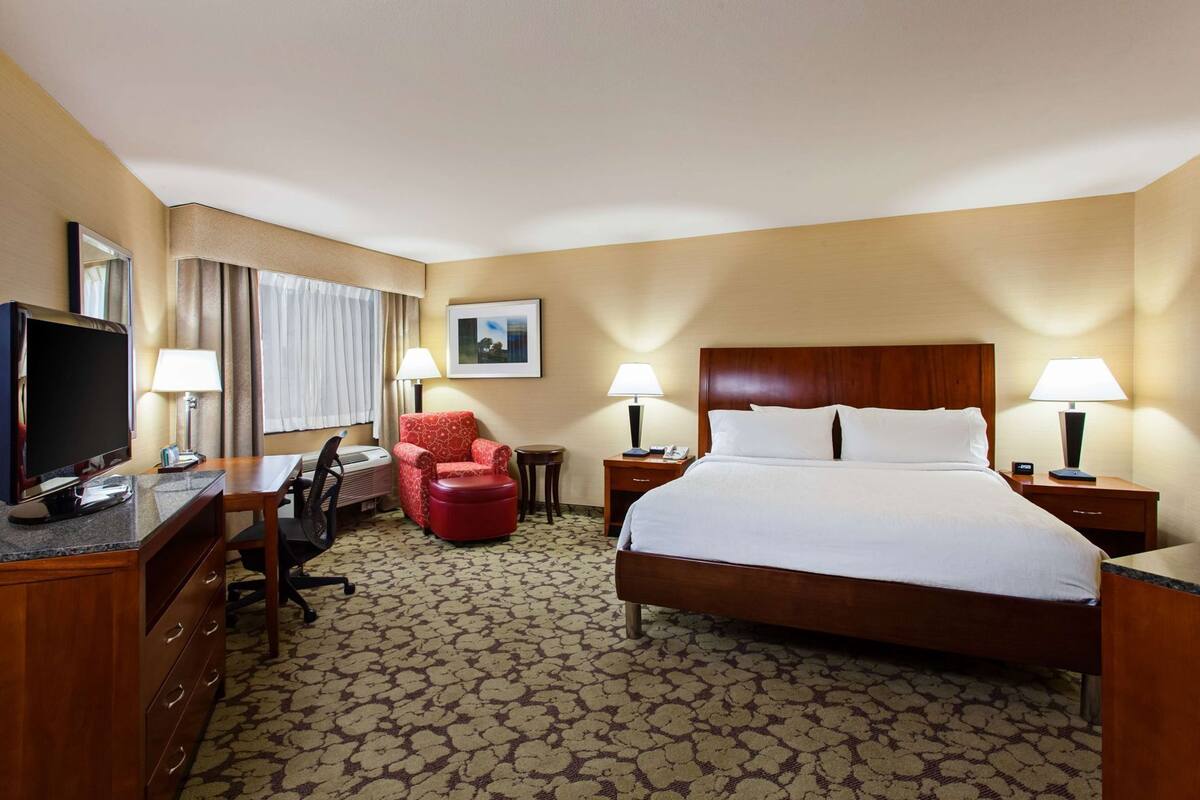 A hotel room with a bed, a desk, a TV, a red armchair, two bedside tables with lamps, and a framed picture on the wall.