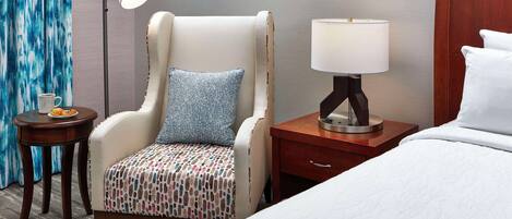 Een hotelkamer met een bed, een stoel met een gestreept kussen, een nachtkastje met een lamp en een klein tafeltje met een kop en bord.