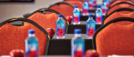 Una fila de habitaciones de hotel con sillas rojas estampadas, mesas con botellas de agua Fiji y manzanas.