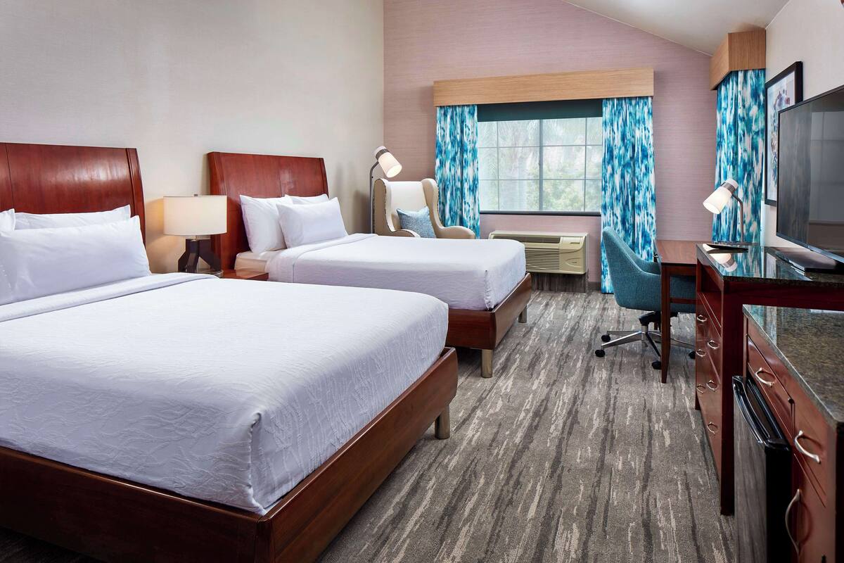 A hotel room with two beds, a desk, a chair, a TV, and a window with curtains.