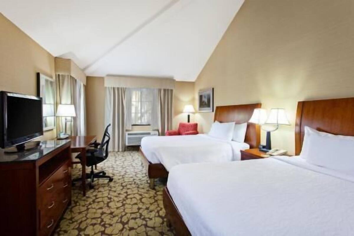 A hotel room with two beds, a desk with a television, a chair, and a lamp.