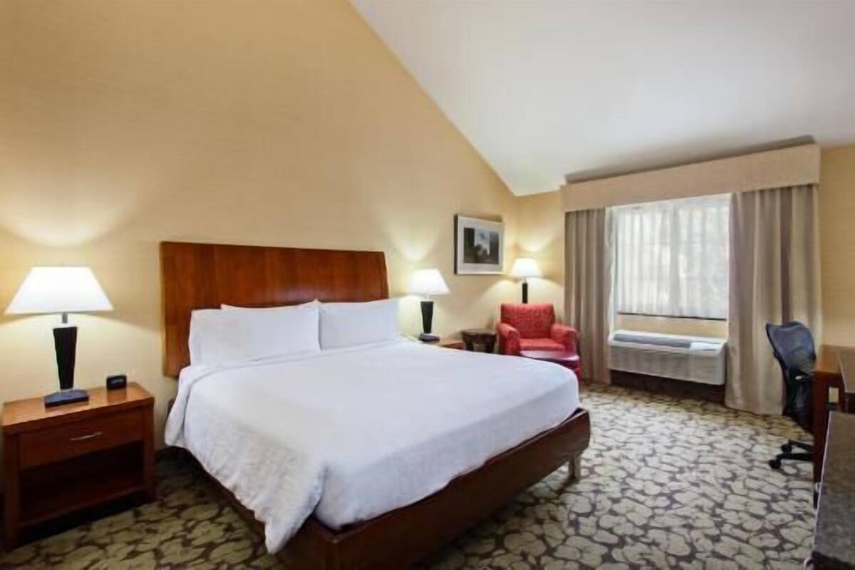 A hotel room with a large bed, two bedside tables with lamps, a red chair, a desk with a chair, and a window with curtains.