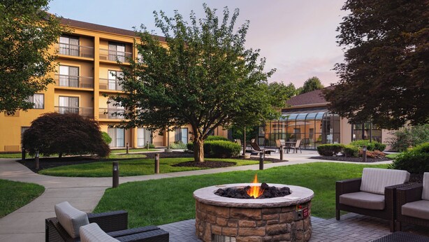 A courtyard with a stone fire pit surrounded by wicker furniture, a pathway, and a building with balconies. - Courtyard by Marriott Fishkill (Fishkill)