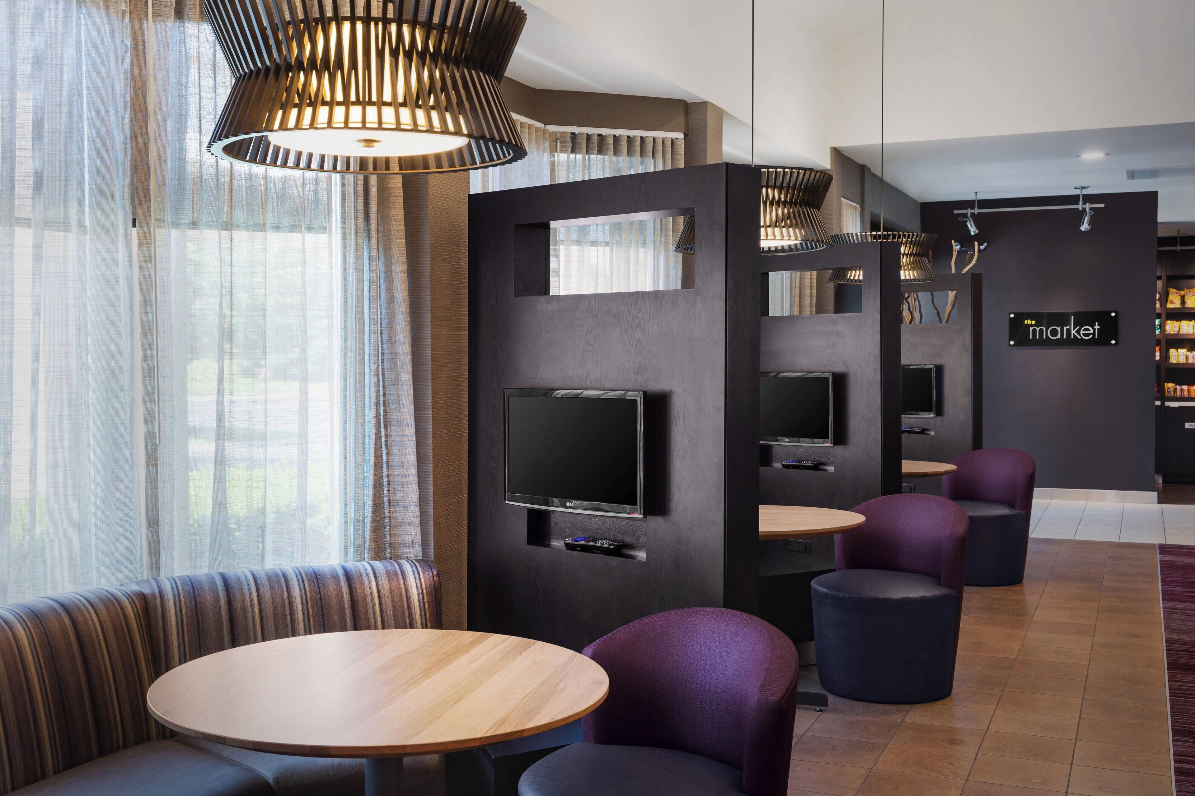 A modern hotel lobby with a seating area featuring a round wooden table and purple chairs, a flat-screen TV mounted on a dark wall, and a sign that reads "market".