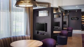 A modern hotel lobby with a seating area featuring a round wooden table and purple chairs, a flat-screen TV mounted on a dark wall, and a sign that reads "market".