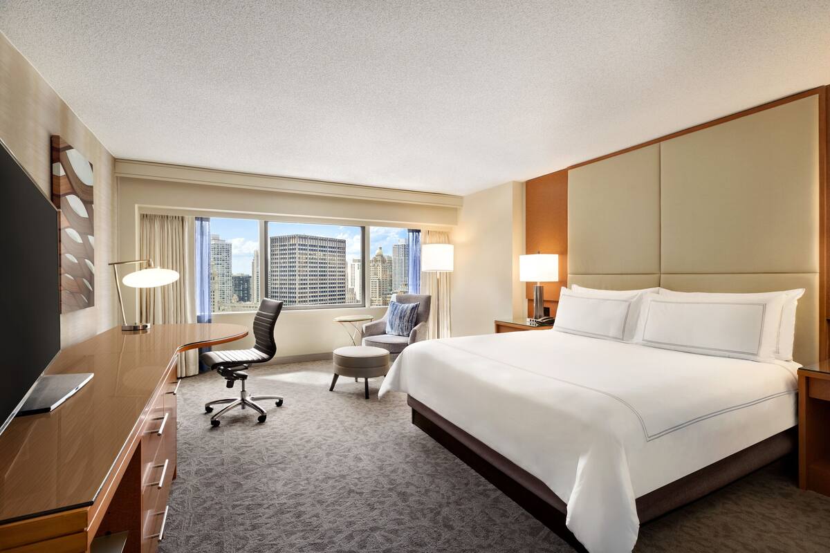 A hotel room with a large bed, a desk with a laptop, a chair, a small ottoman, and a view of the city skyline.