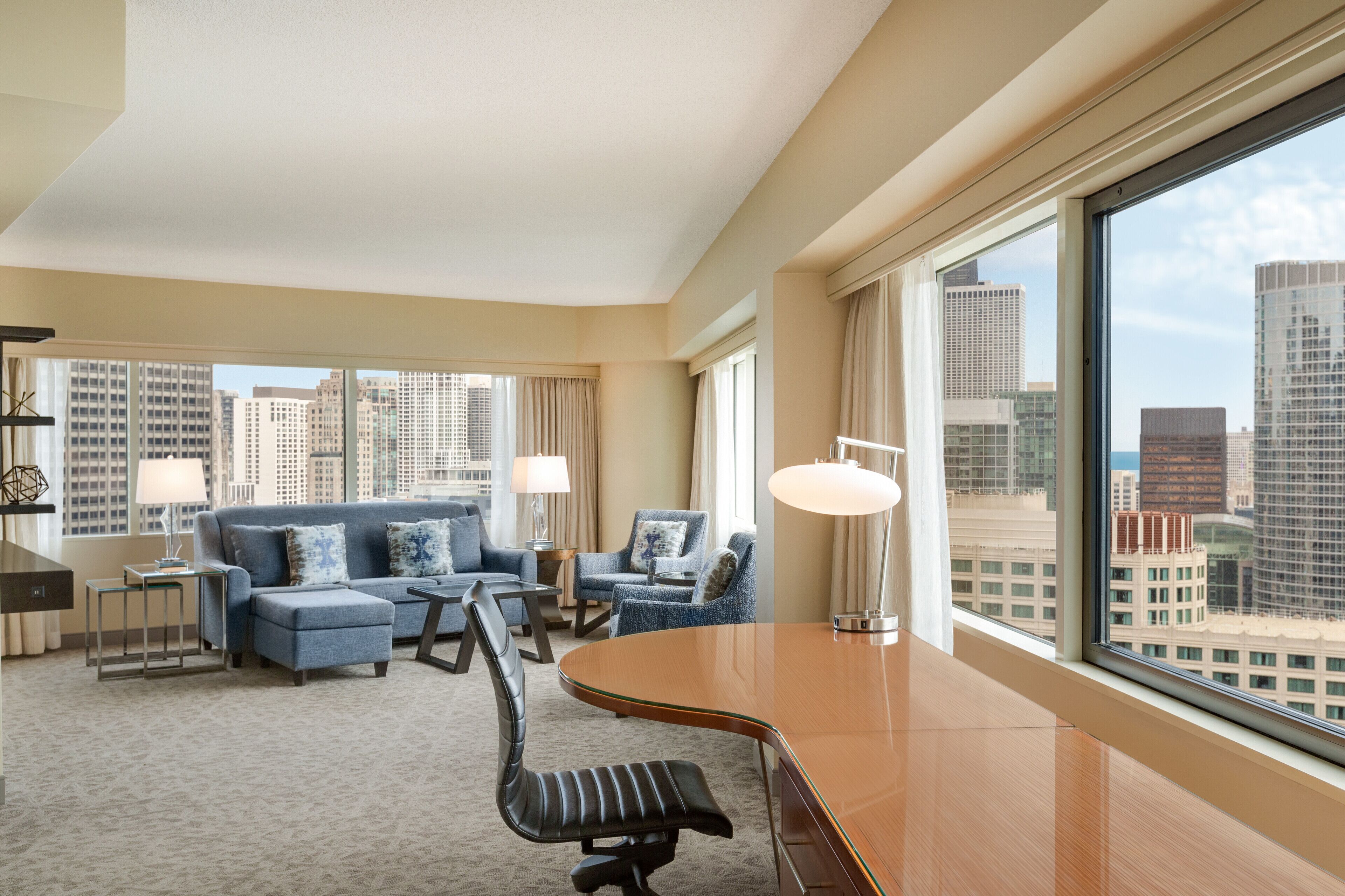 A hotel room with a large window, a sofa, two armchairs, a desk, and a chair.
