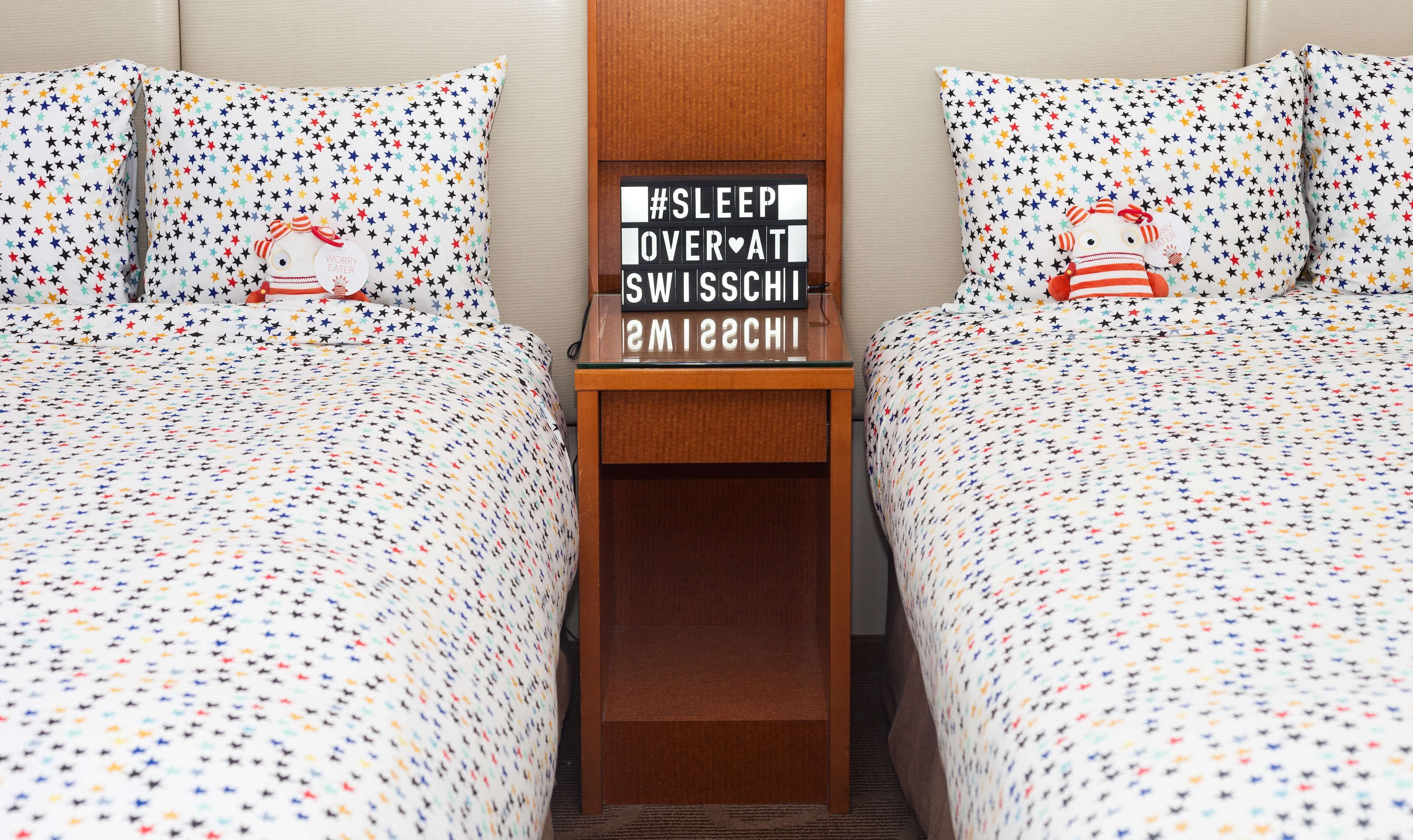 Habitación de hotel con dos camas, cada una con una colcha blanca con un estampado de estrellas de colores. Hay una mesita de noche de madera entre las camas con un cartel que dice "#SLEEP OVER AT SWISSCHISCH".