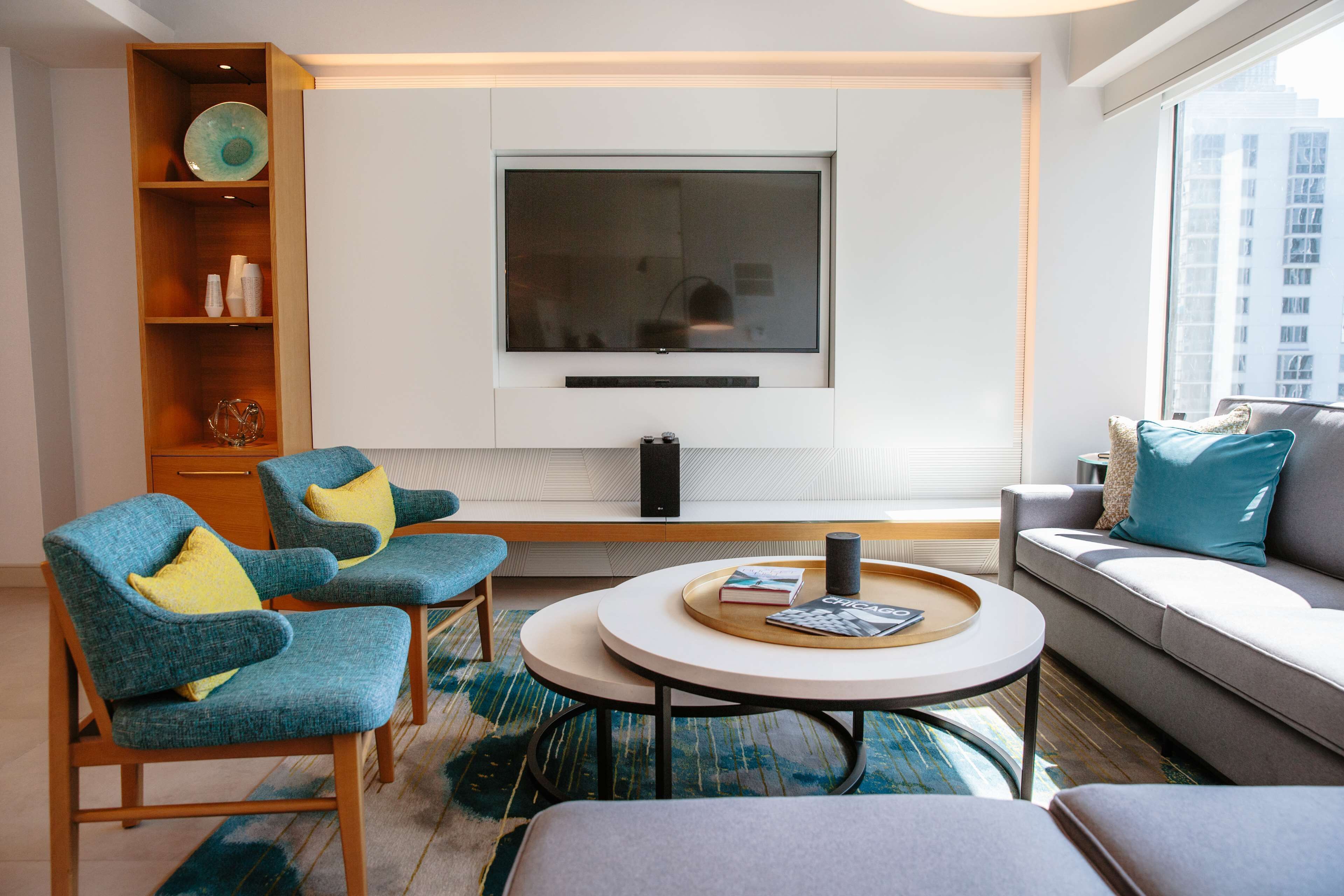 Una sala de estar moderna con un televisor de pantalla plana en la pared, dos sillones azules con cojines amarillos, un sofá gris con cojines verde azulado y una mesa de centro redonda con libros y una bandeja.
