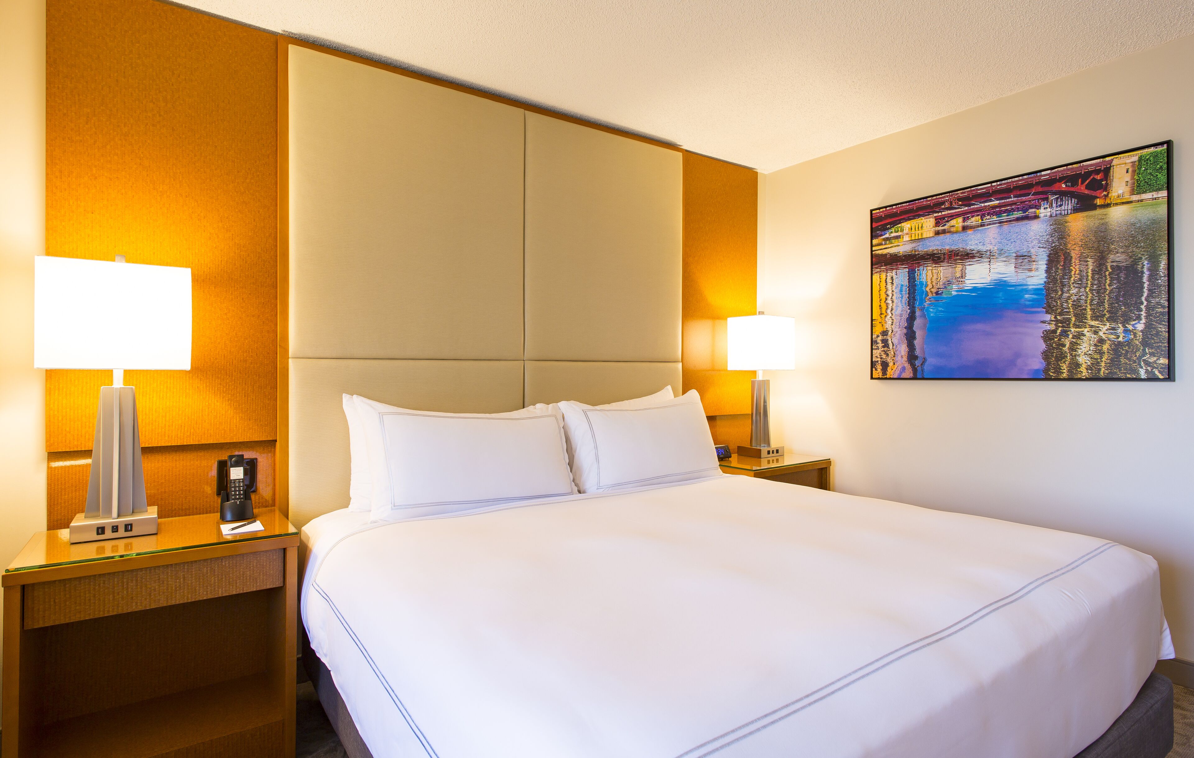 Habitación de hotel con una cama grande, dos mesitas de noche con lámparas, un teléfono y un cuadro en la pared.