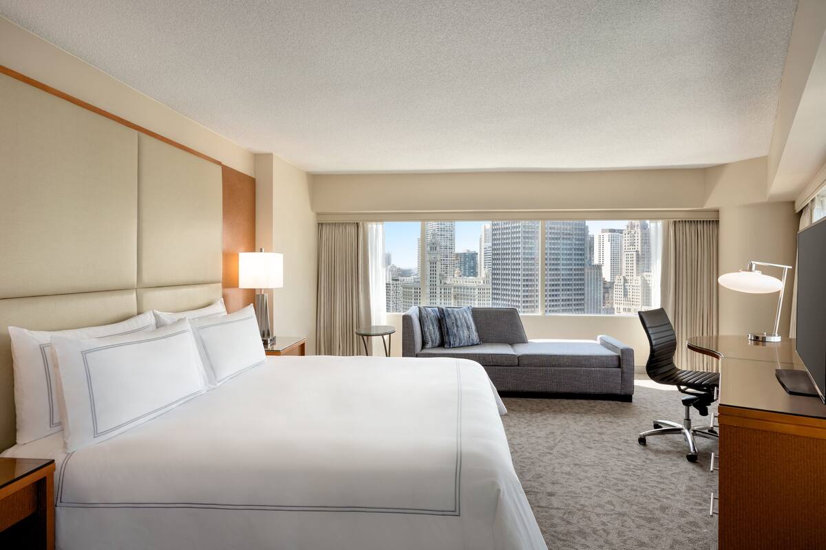 A hotel room with a large bed, a sofa, a desk with a chair, and a TV.