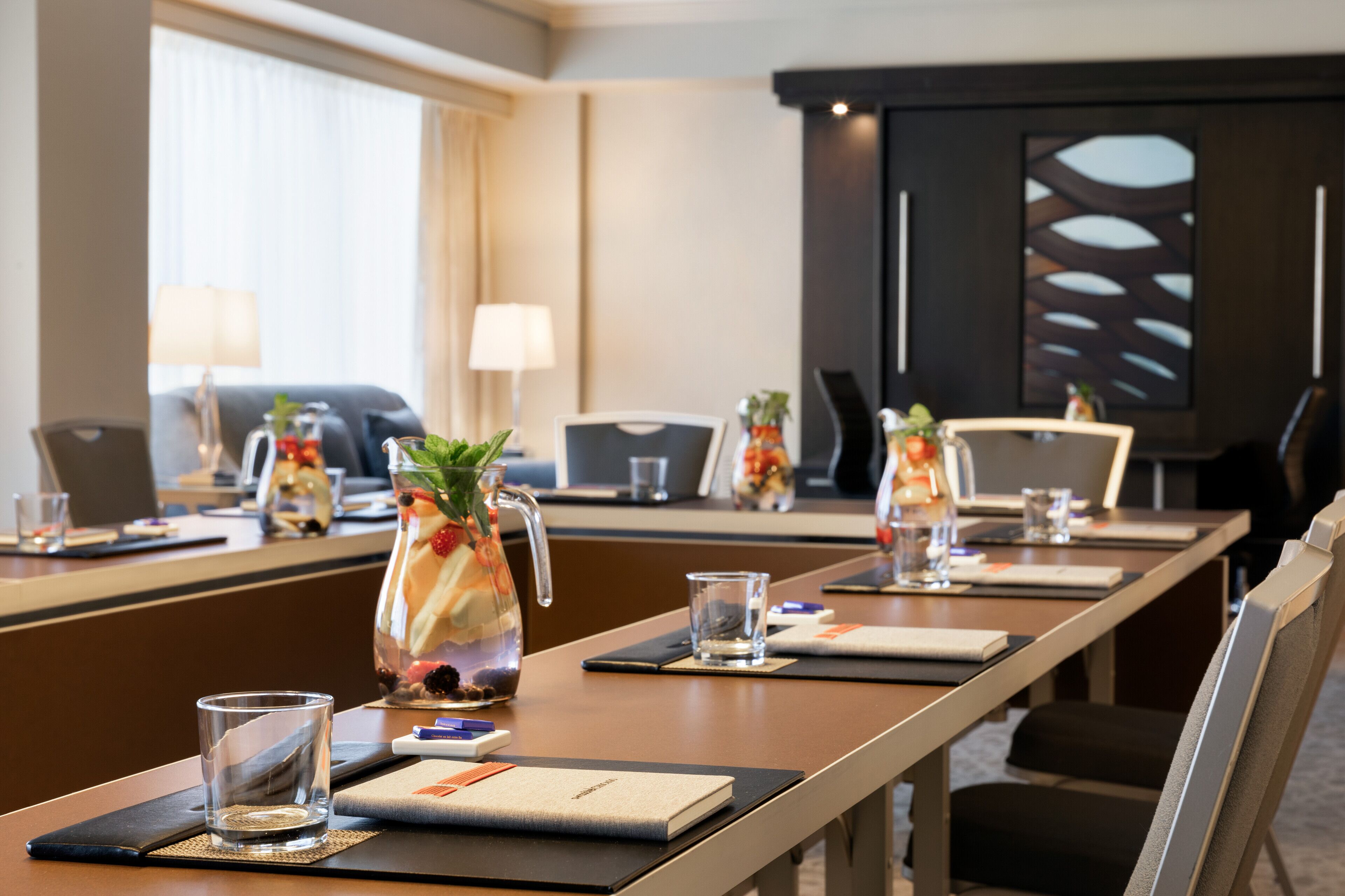 A conference room with a long table, chairs, a projector screen, and a decorative pitcher with fruits and a plant.