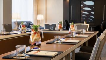 A conference room with a long table, chairs, a projector screen, and a decorative pitcher with fruits and a plant.