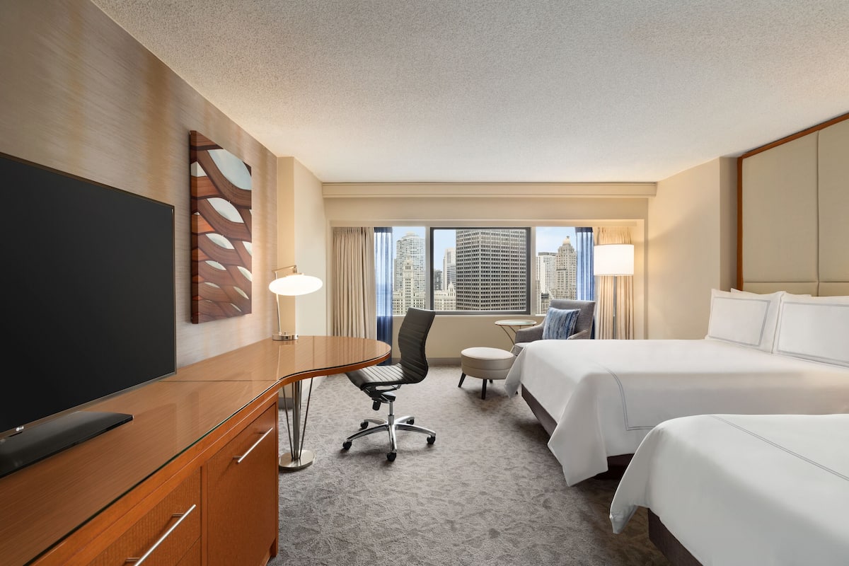 A hotel room with a large flat-screen TV, a wooden desk, a modern chair, two beds, a small round table, and a view of the city skyline through large windows.
