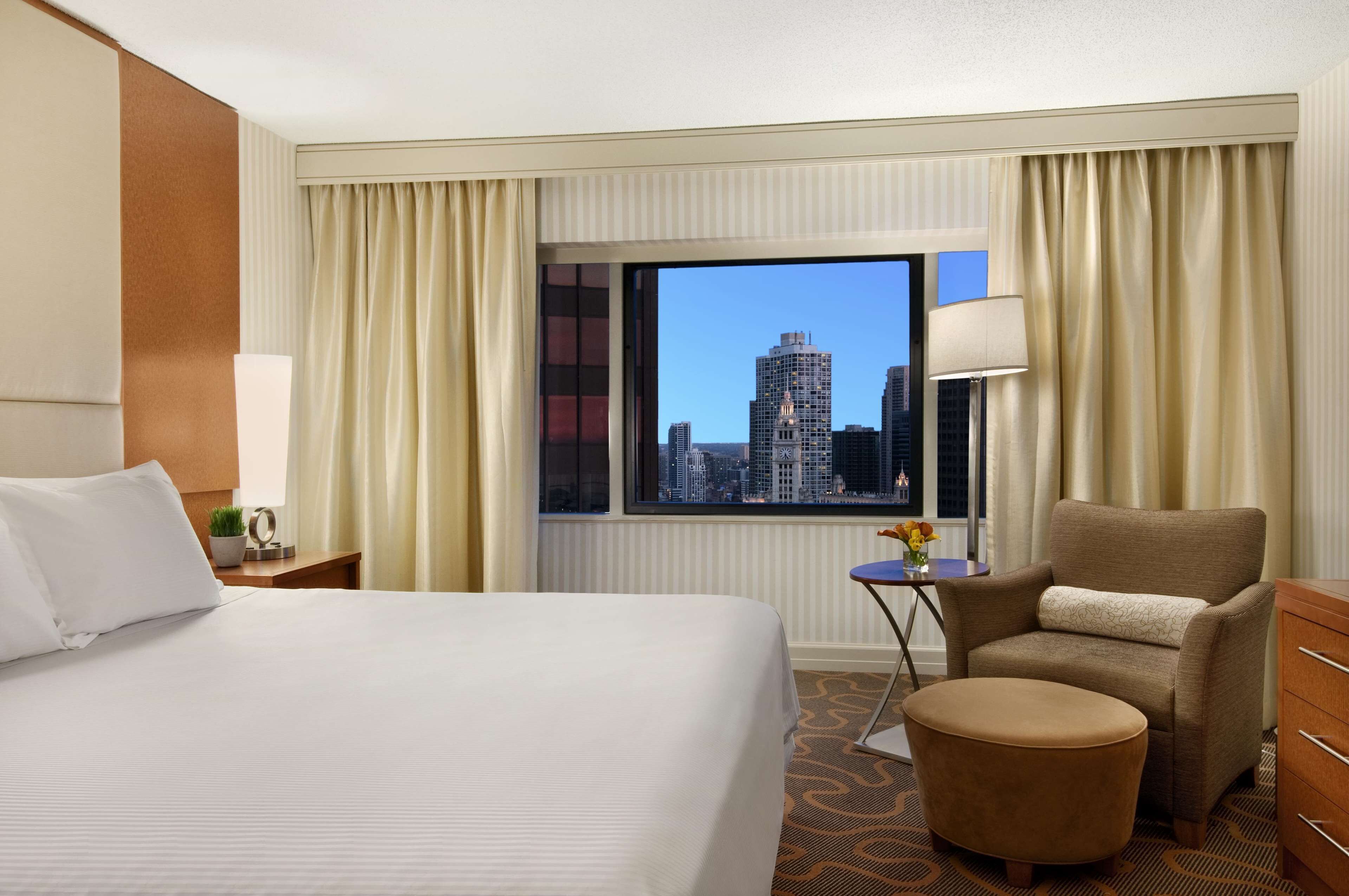 Quarto de hotel com uma cama grande, uma cadeira, uma mesinha com abajur e vista para a paisagem urbana através de uma janela.