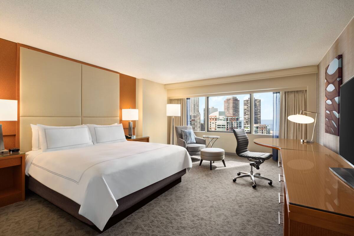 A hotel room with a large bed, a desk, two chairs, a TV, and a view of the city.