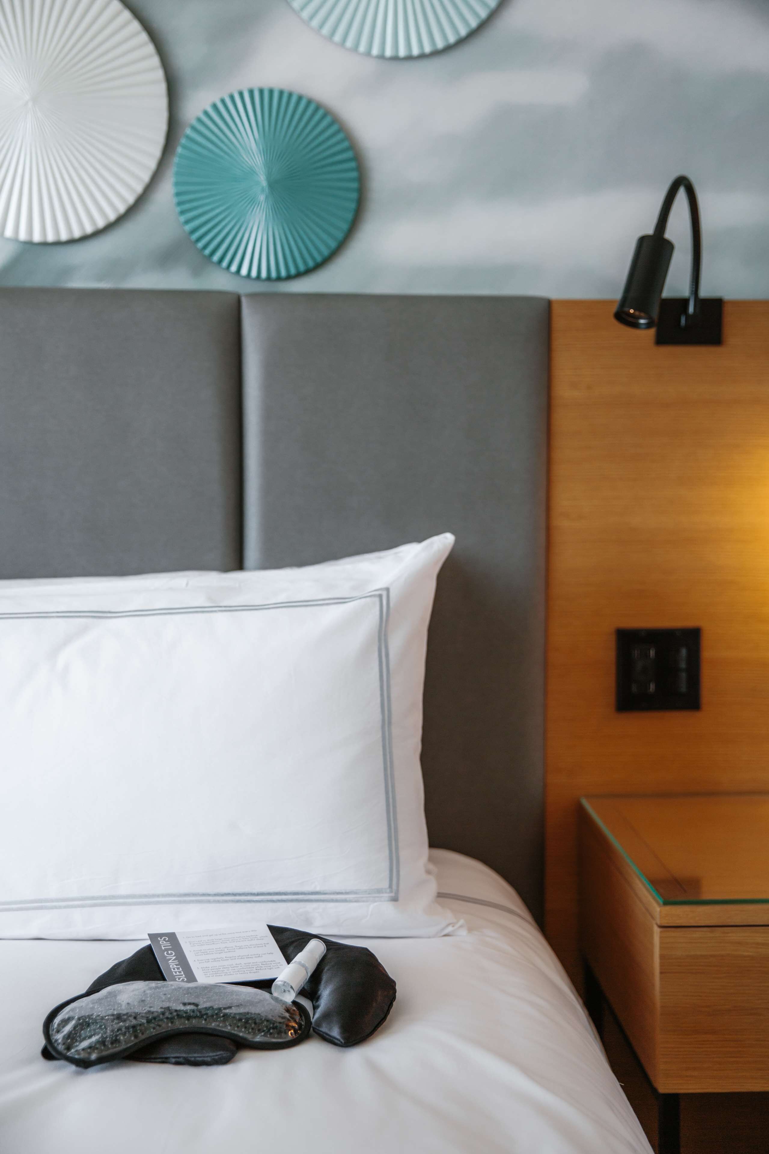 A neatly made bed with white bedding, a black pillow, and a black eye mask. There is a wooden nightstand with a lamp and a small table beside the bed.