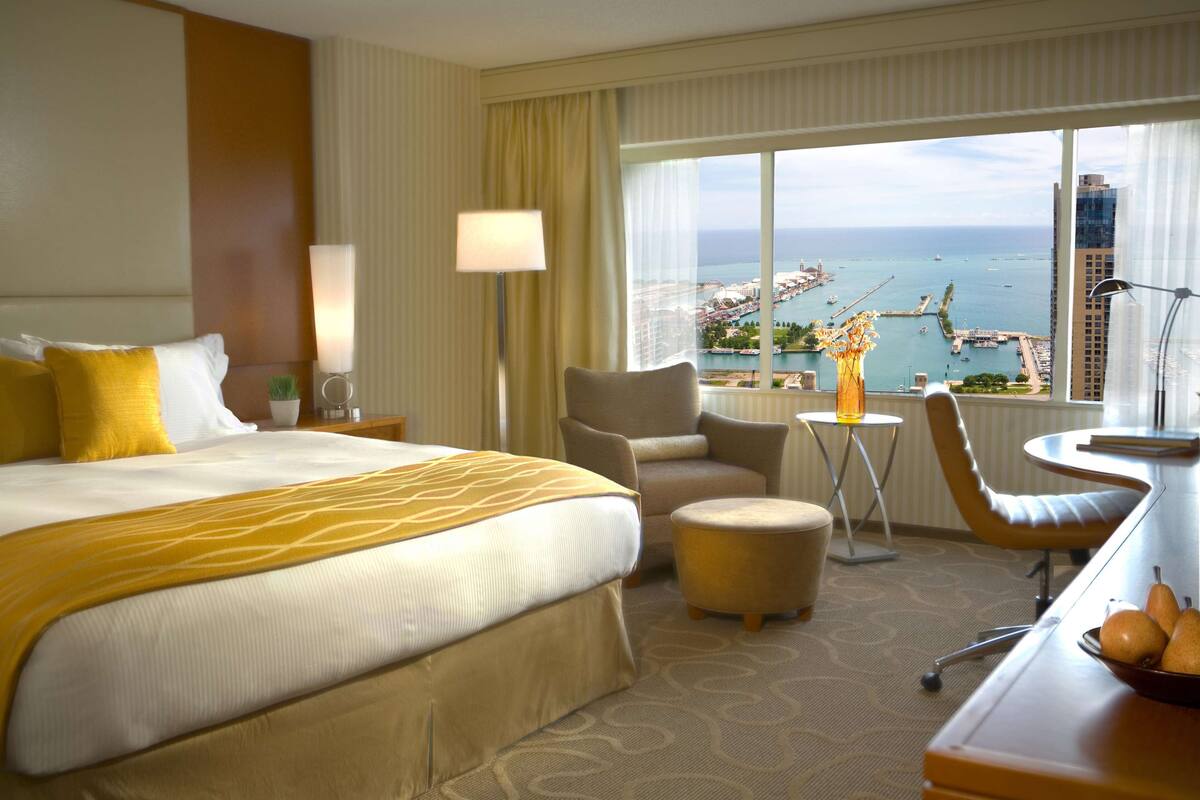 A hotel room with a large bed, a desk with a chair, a small table with a lamp, and a view of the ocean and cityscape.