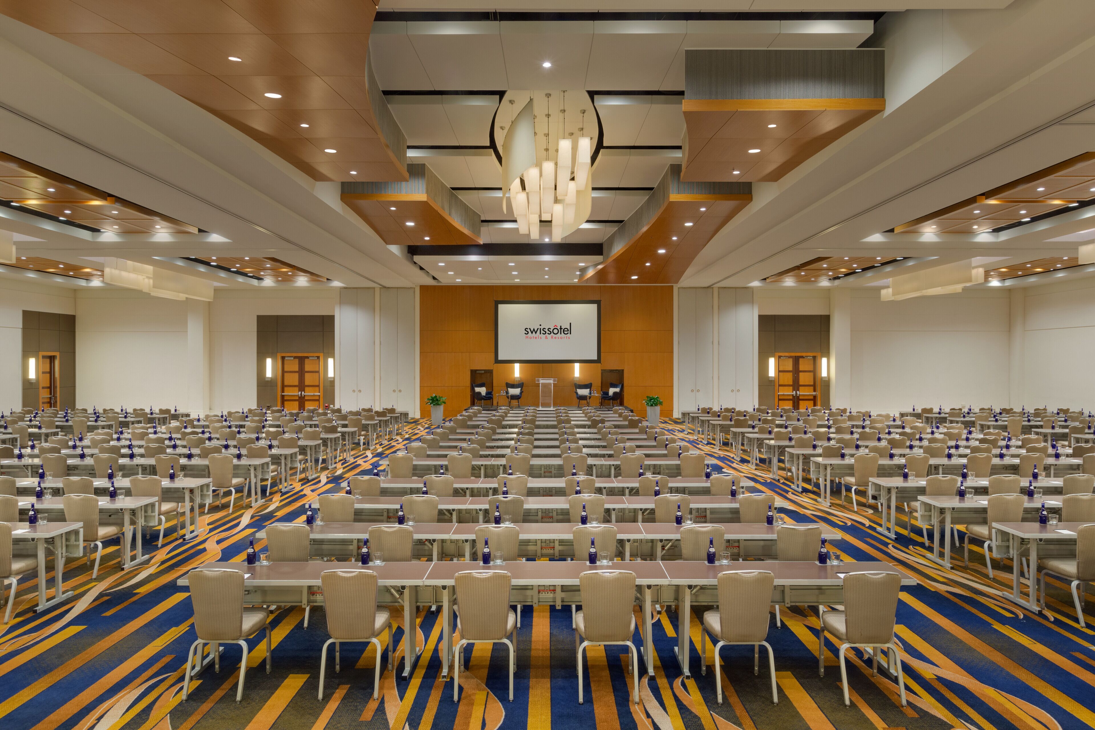 A large conference room with rows of tables and chairs, a central screen, and a modern chandelier.