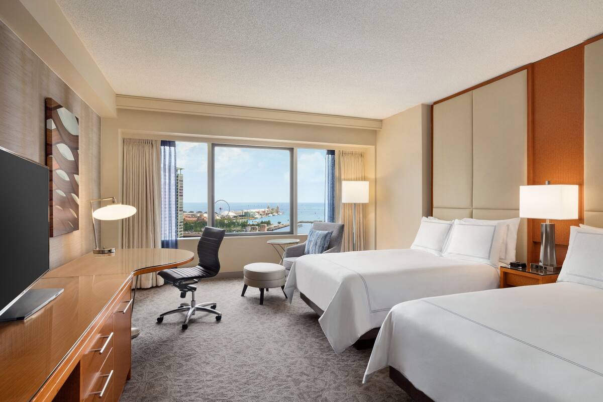 A hotel room with two beds, a desk, a chair, a TV, and a window with a view of the city.