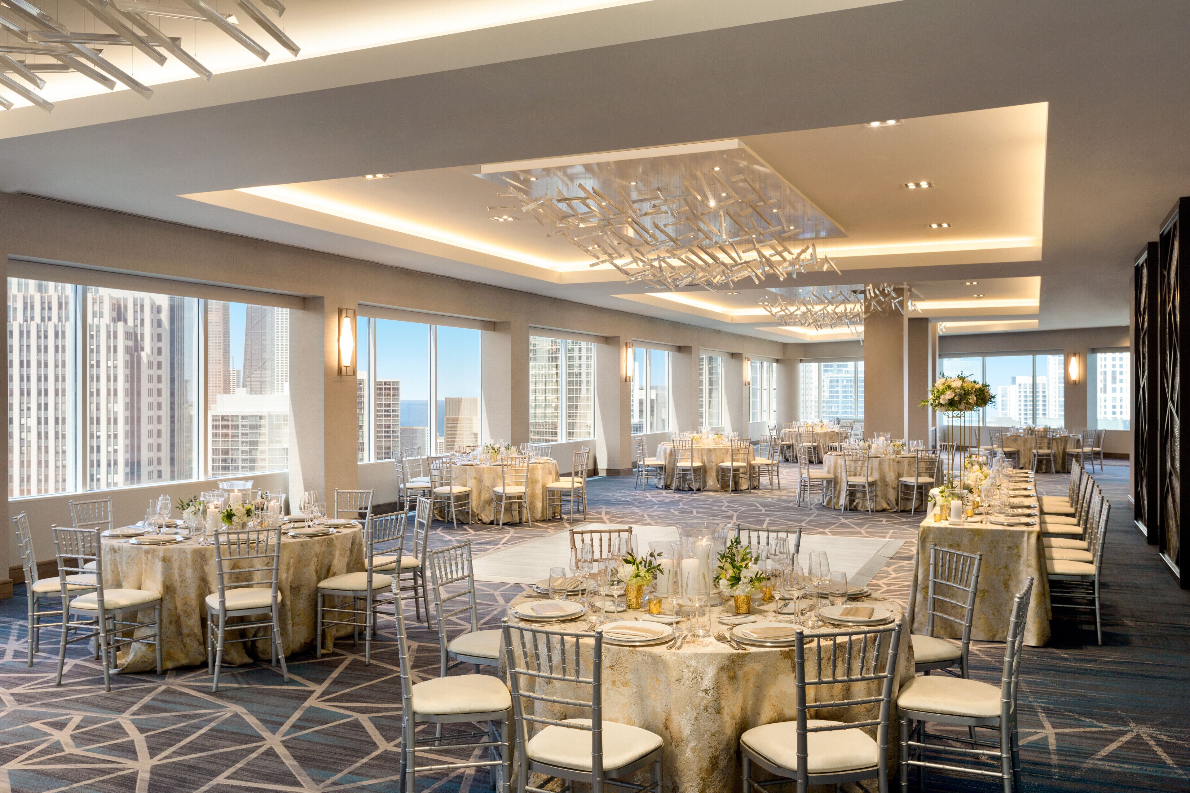 Un amplio salón de banquetes con varias mesas redondas preparadas para cenar, ventanas grandes que ofrecen vistas de la ciudad y un diseño de techo moderno.