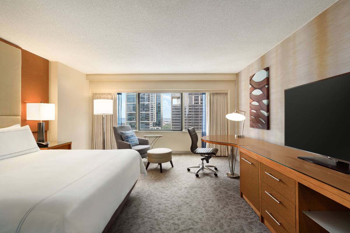 A hotel room with a large bed, a desk with a flat-screen TV, a chair, a small table, and a view of tall buildings through the window.