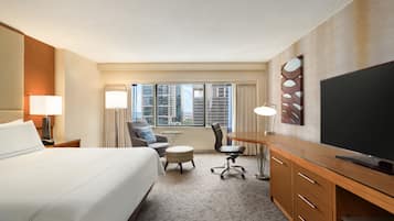Ein Hotelzimmer mit einem großen Bett, einem Schreibtisch mit Flachbildfernseher, einem Sessel, einem kleinen Tisch und Blick auf hohe Gebäude durch das Fenster.