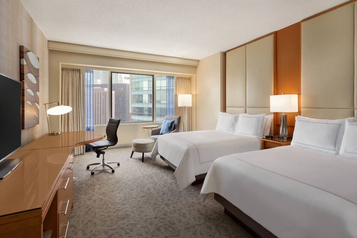A hotel room with two beds, a desk with a flat-screen TV, a chair, a small round table, and a large window with curtains.