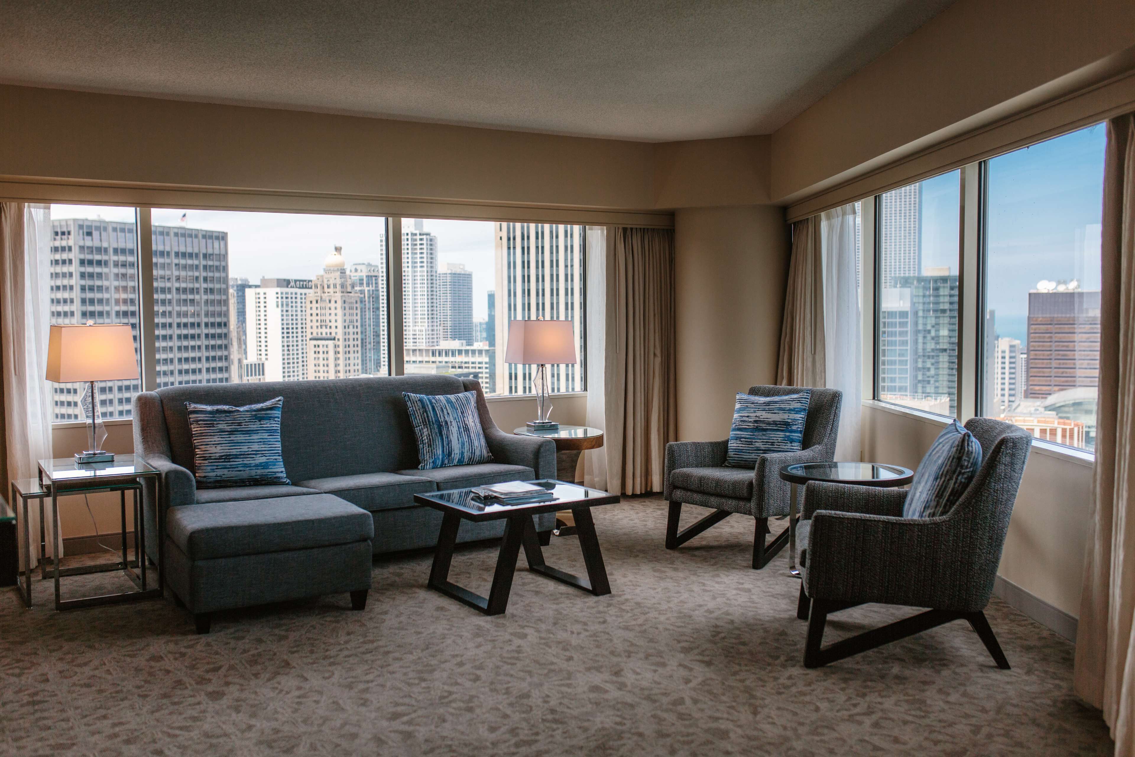 A hotel room with a large window, a sofa, a chair, a small table, and a lamp.