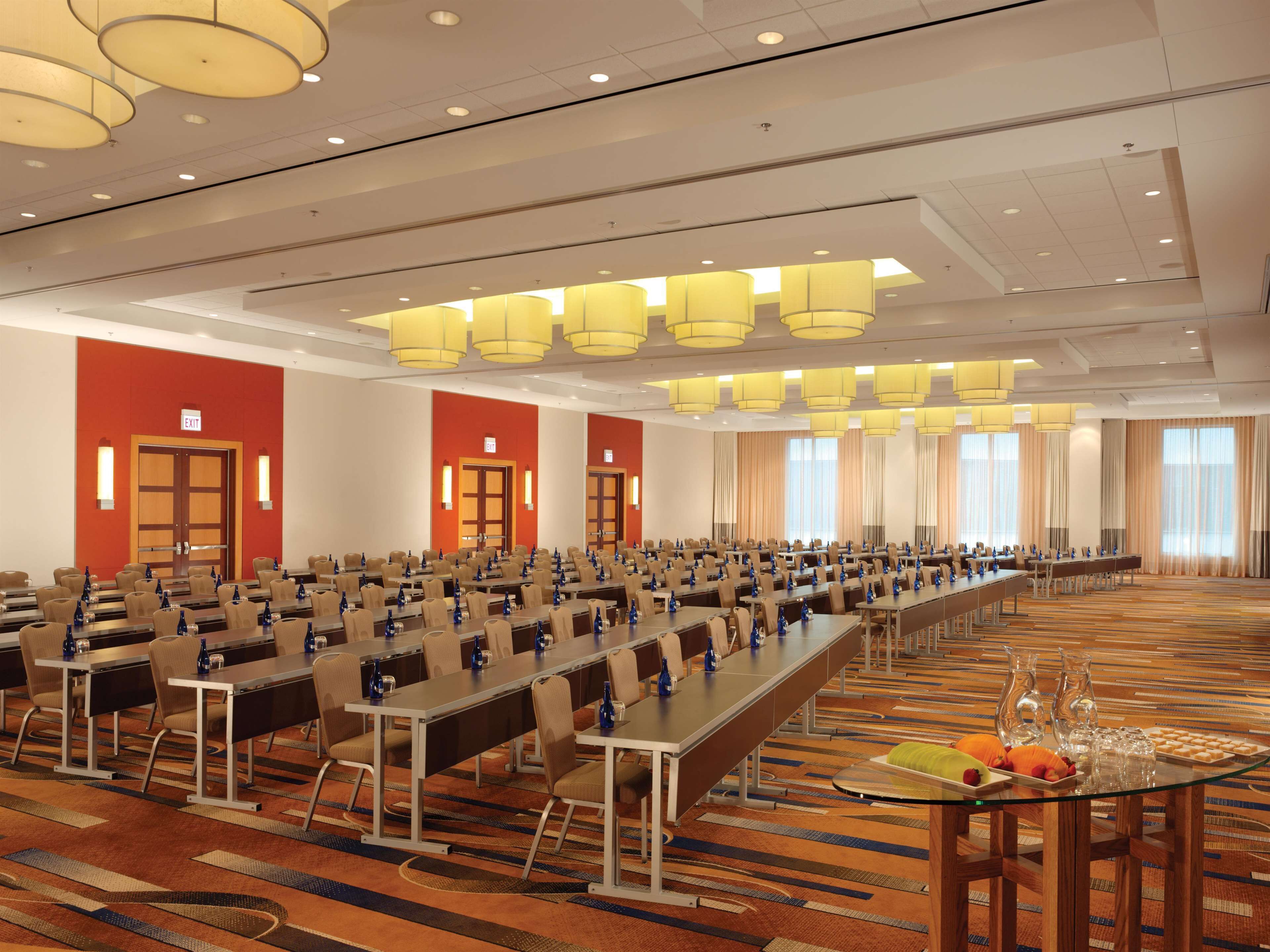 Un amplio salón de conferencias con filas de mesas y sillas, equipado con botellas de agua y una mesa de buffet con refrigerios.