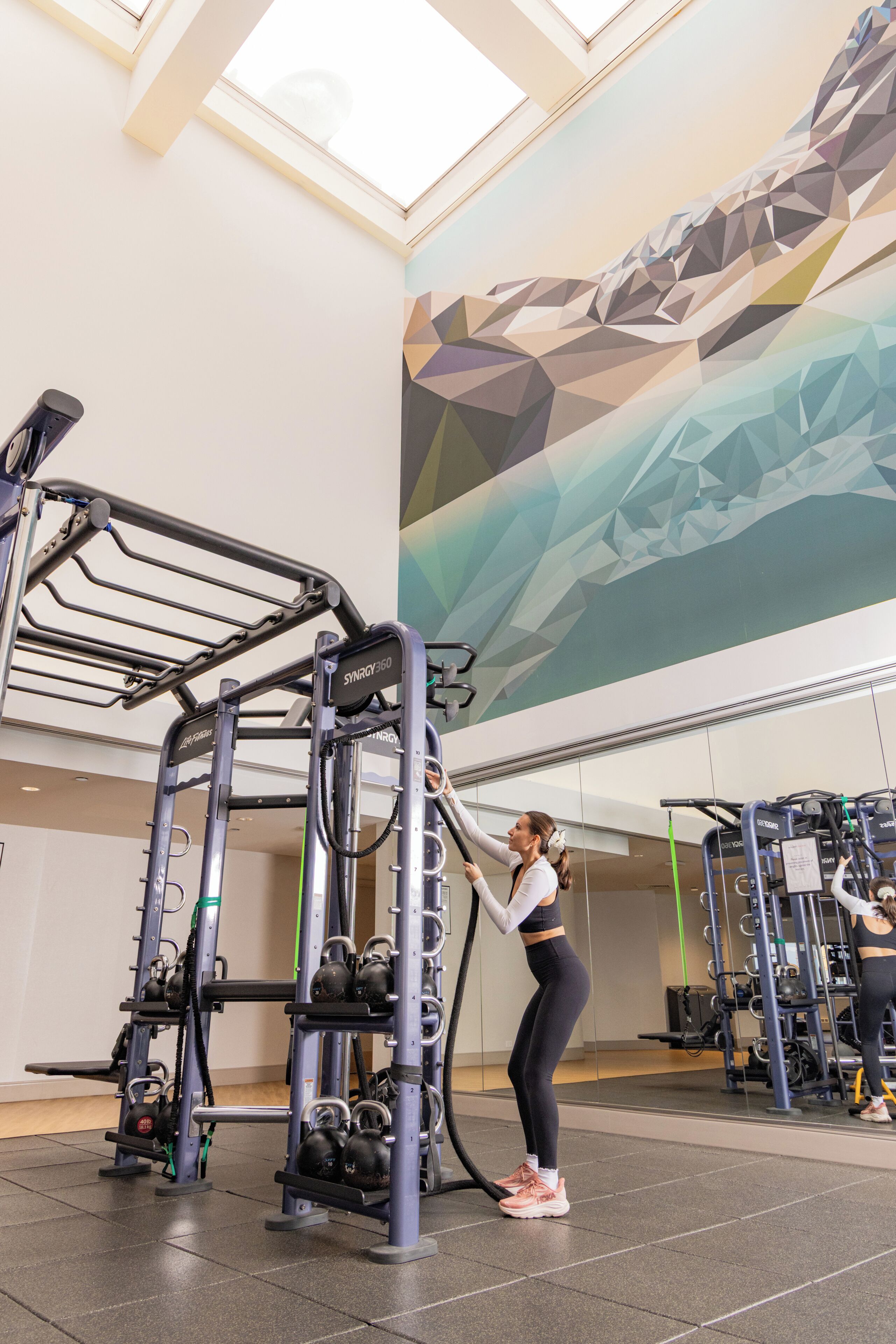 A gym with exercise equipment, a high ceiling, and a large abstract mural on the wall.