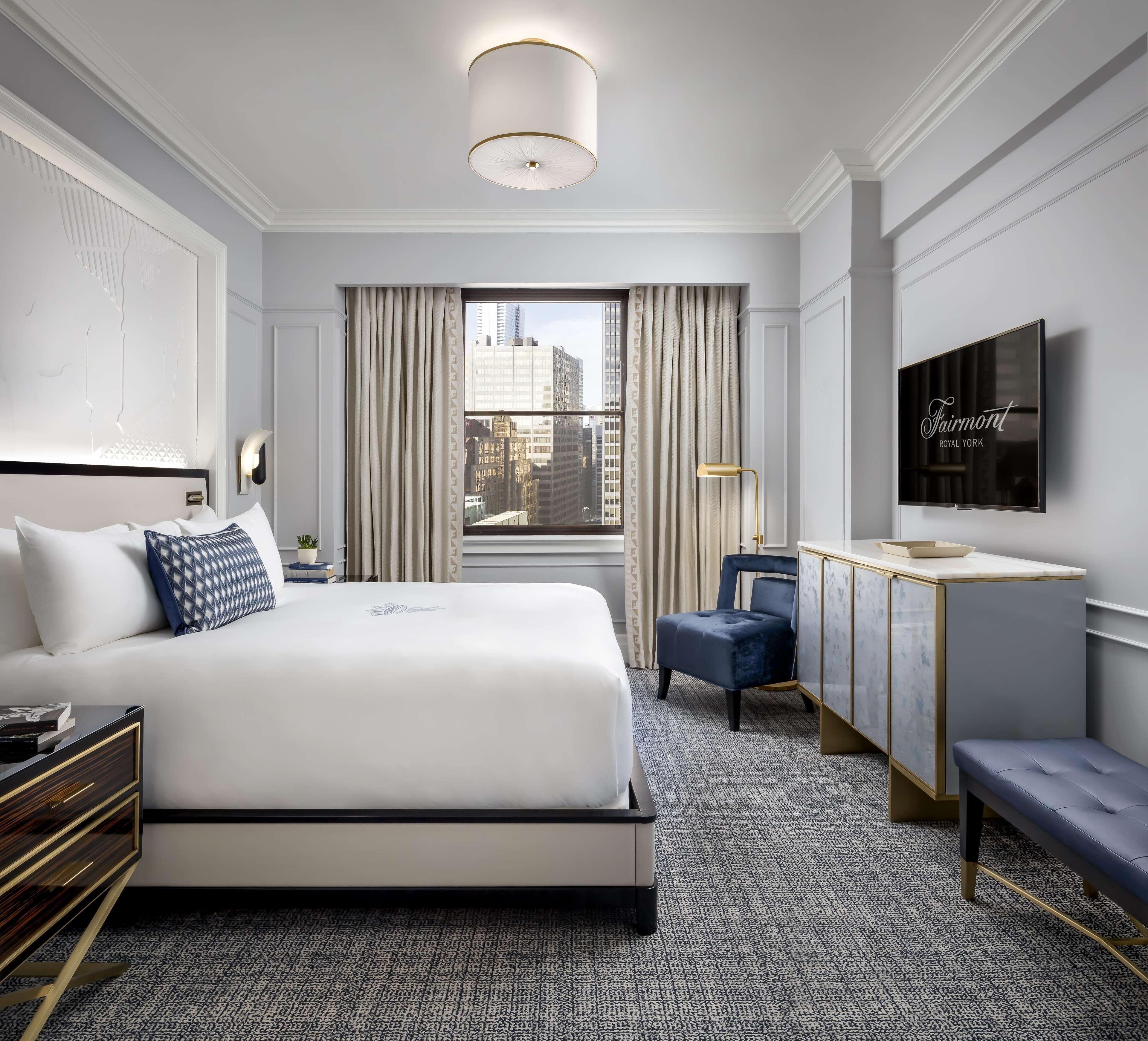 A hotel room with a large bed, a desk with a chair, a television, and a view of the city through the window.