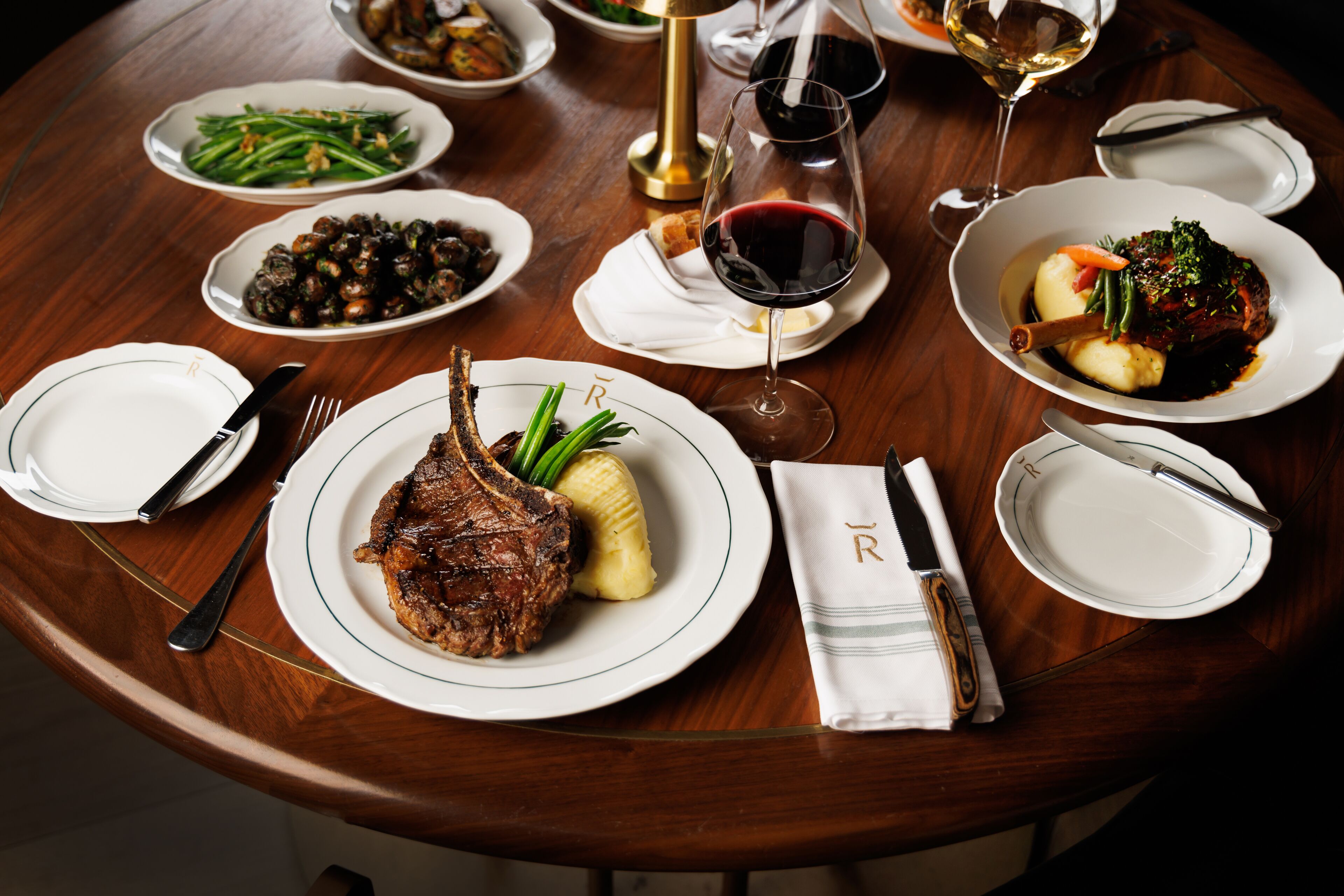 A round wooden table with various dishes, including a steak, vegetables, and a glass of red wine.