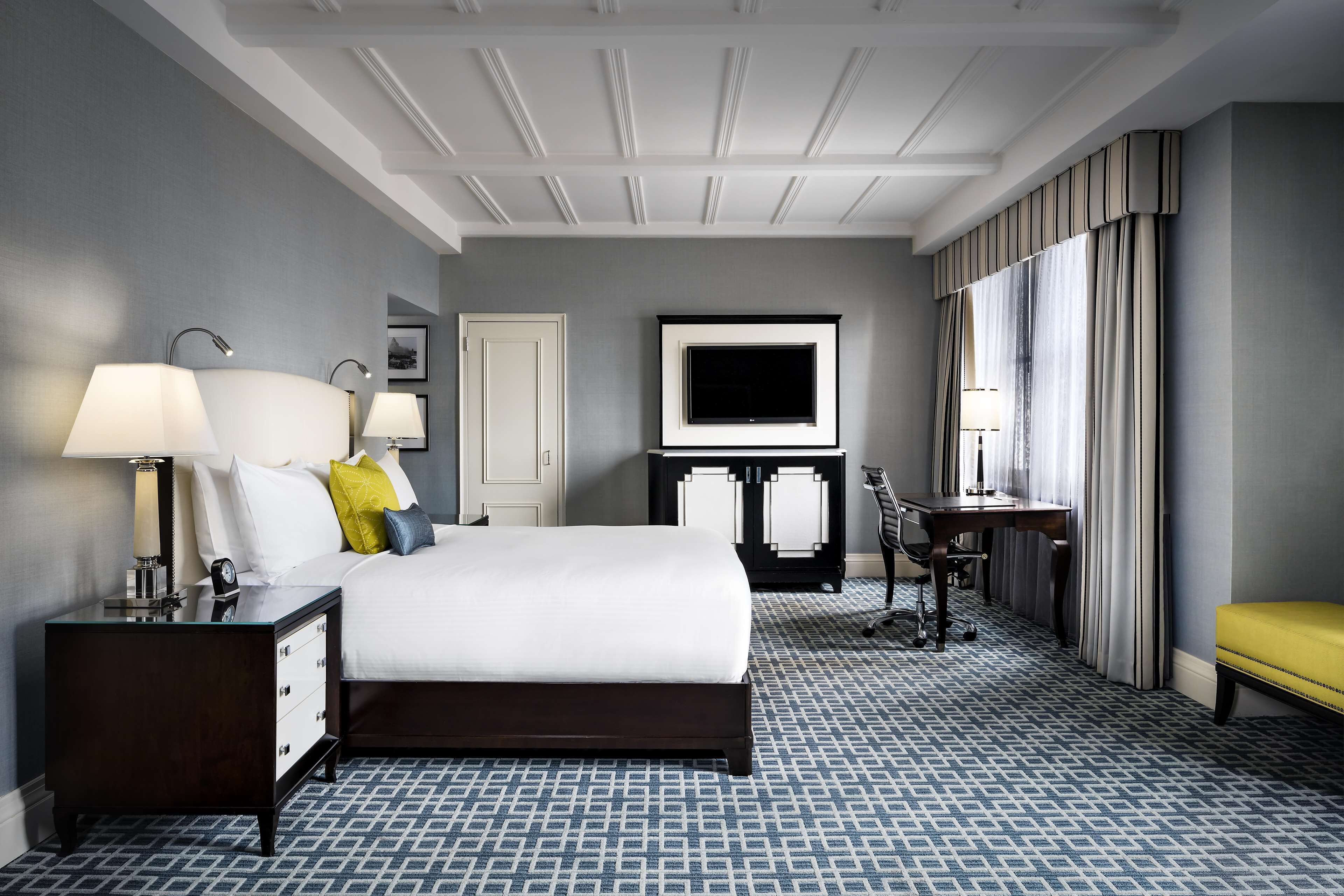 A hotel room with a large bed, bedside tables with lamps, a desk, a chair, a television, and a window with curtains.