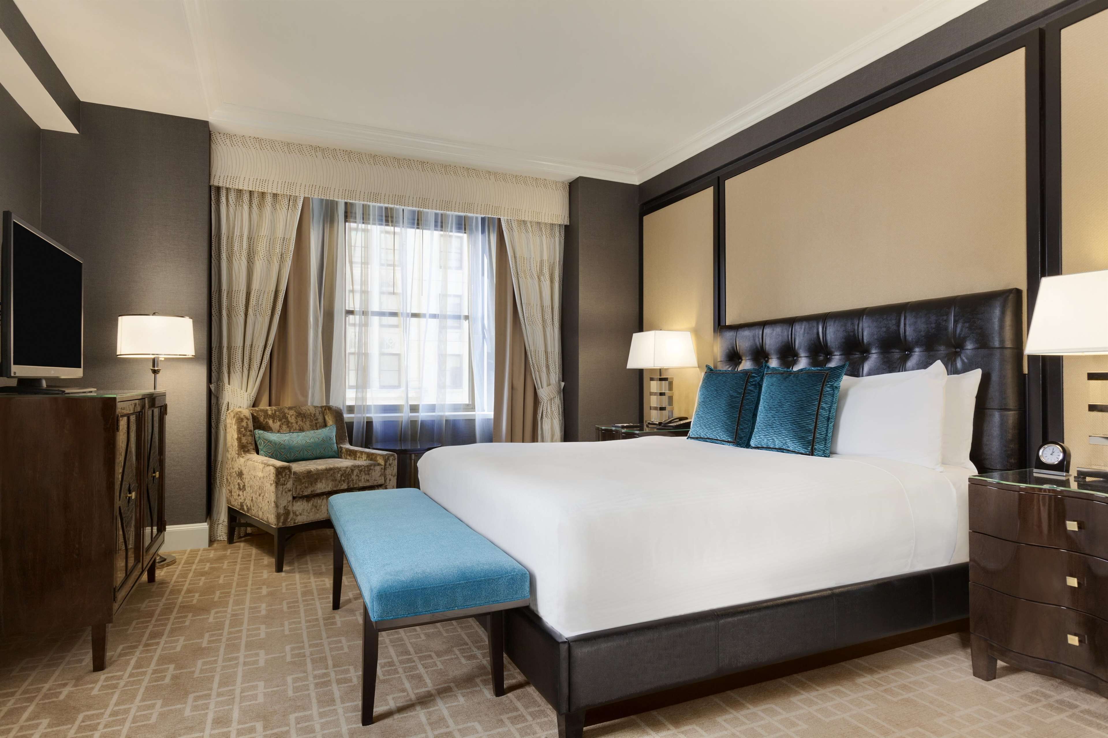 A hotel room with a large bed, two bedside tables, a chair, a bench, a TV, and a window with curtains.