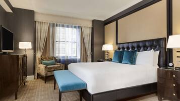Habitación de hotel con una cama grande, dos mesitas de noche, una silla, un banco, un televisor y una ventana con cortinas.