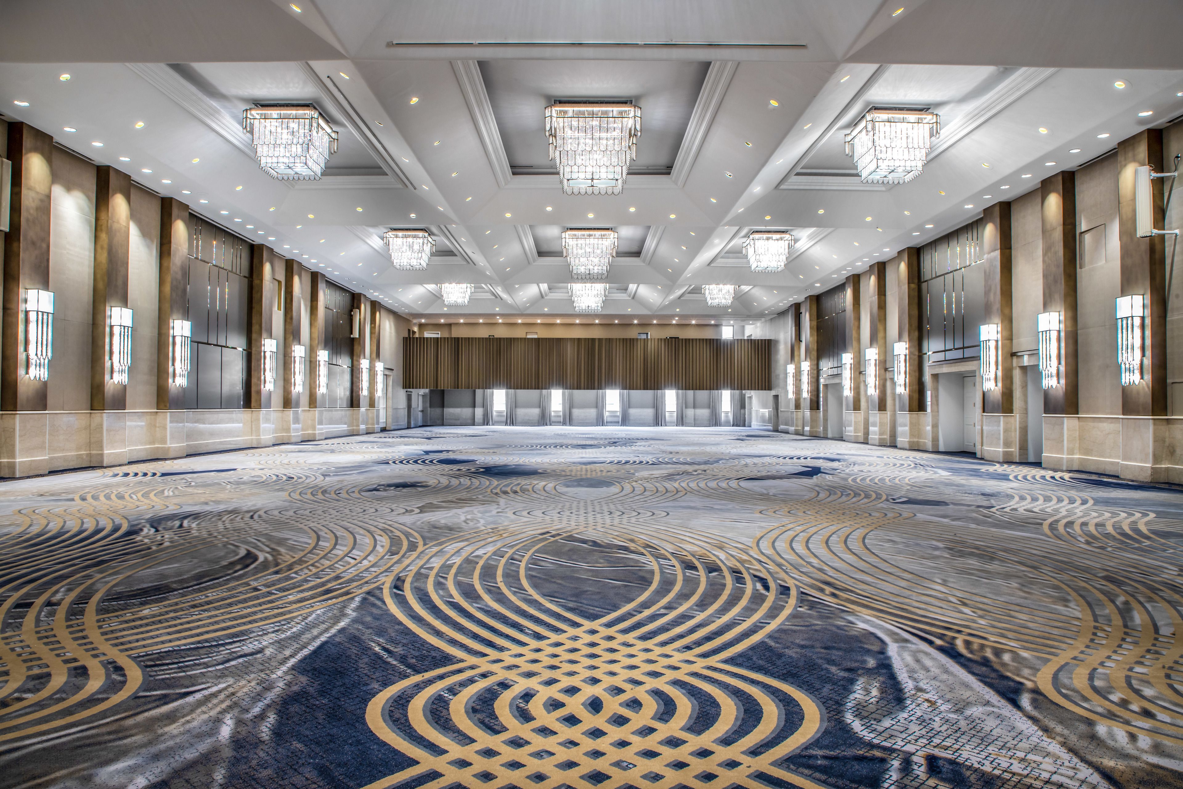 A spacious conference room with a patterned carpet, large chandeliers, and a long row of windows.