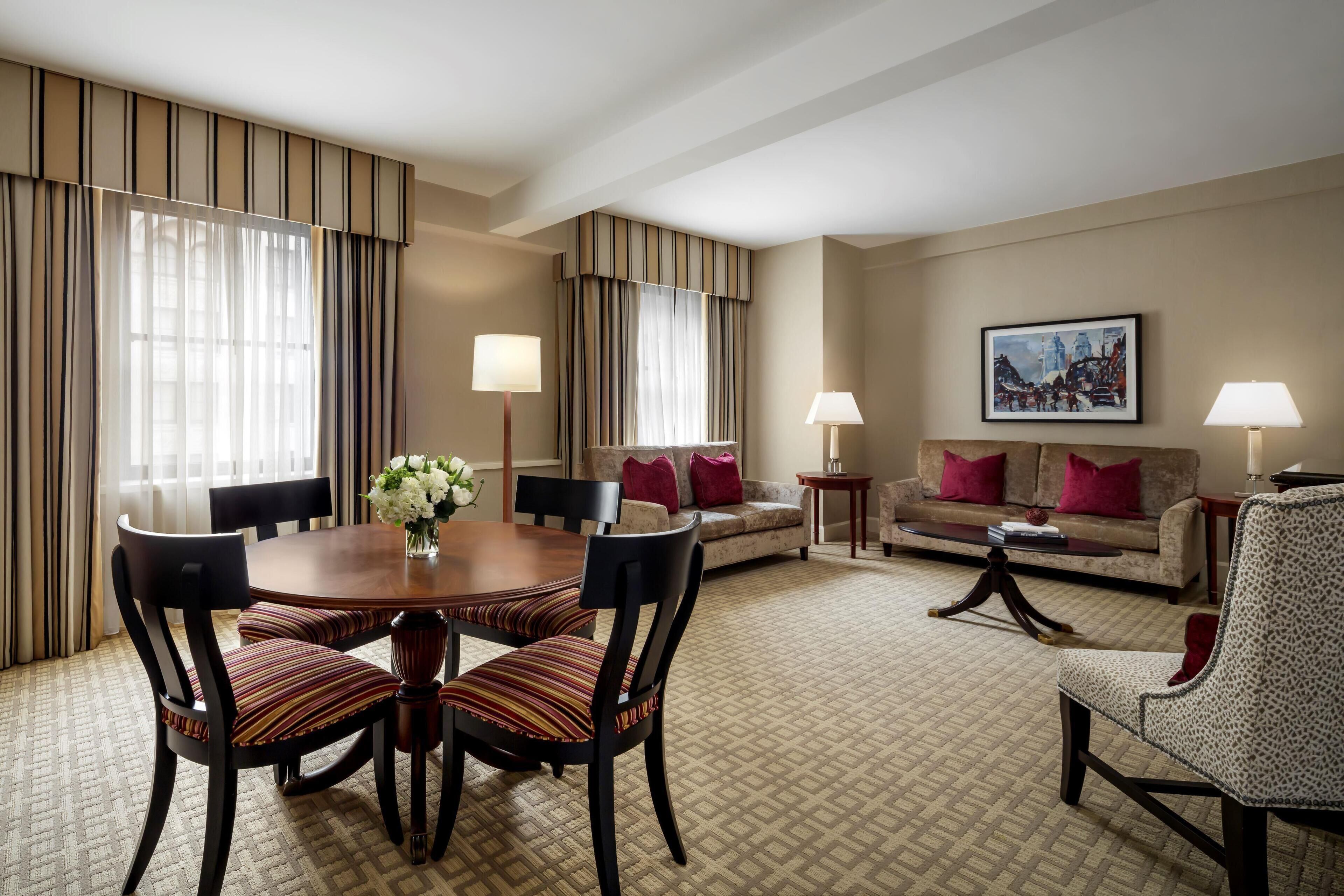 A hotel room with a dining table, chairs, a sofa, a coffee table, and a painting on the wall.