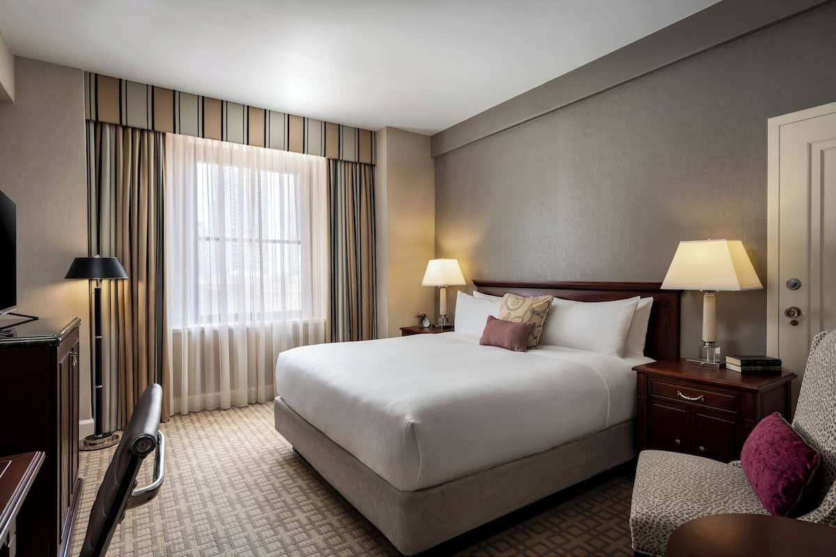 Una habitación de hotel con una cama grande, dos mesitas de noche con lámparas, una silla, un escritorio y un televisor.