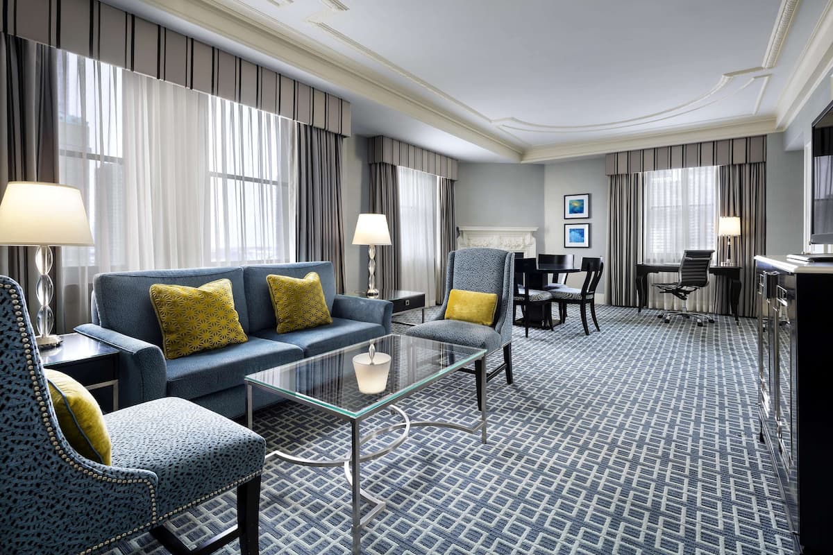 Habitación de hotel con alfombra azul estampada, un sofá, una silla, una mesa de centro de vidrio y un escritorio con una silla.