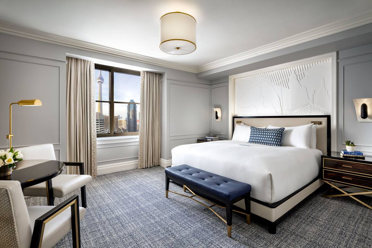 Habitación de hotel con una cama grande, un escritorio con una silla, un banco y vistas a un paisaje urbano a través de una ventana.
