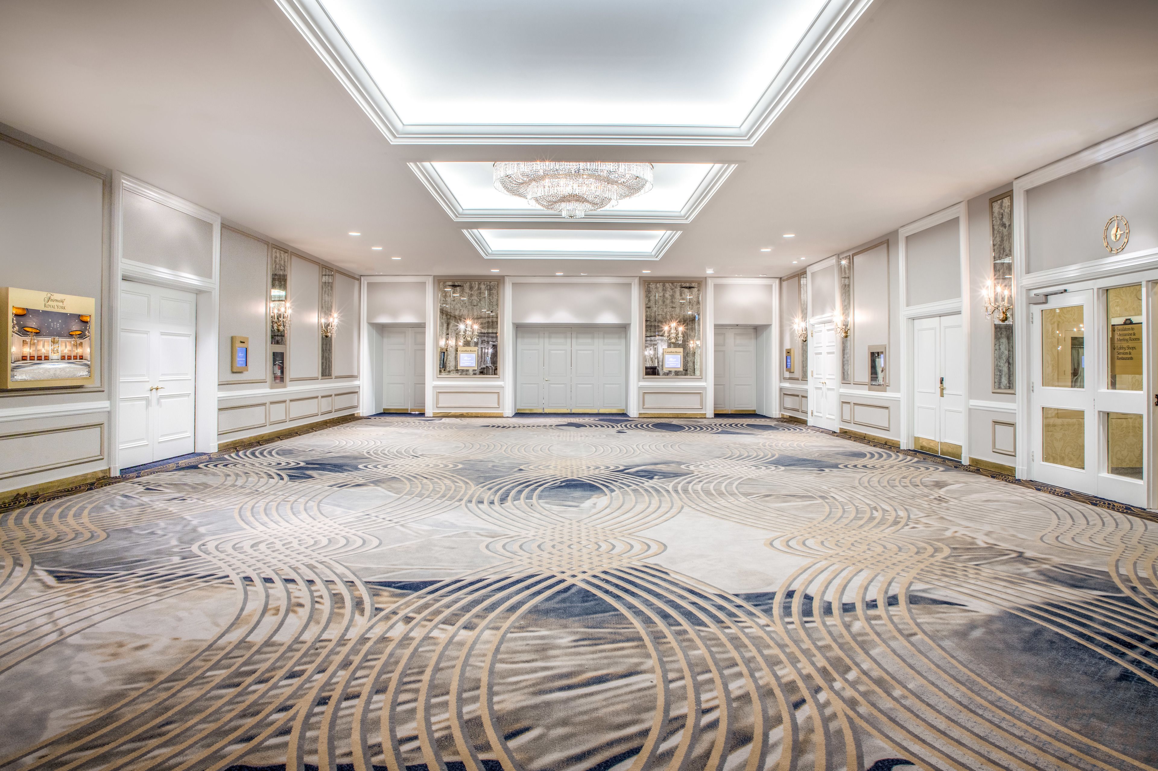 A spacious hotel lobby with a large, patterned carpet, a chandelier, and a modern ceiling design.