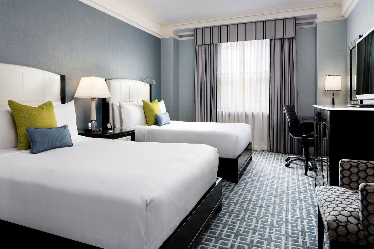 Habitación de hotel con dos camas, un escritorio con una silla y un ventanal con cortinas.
