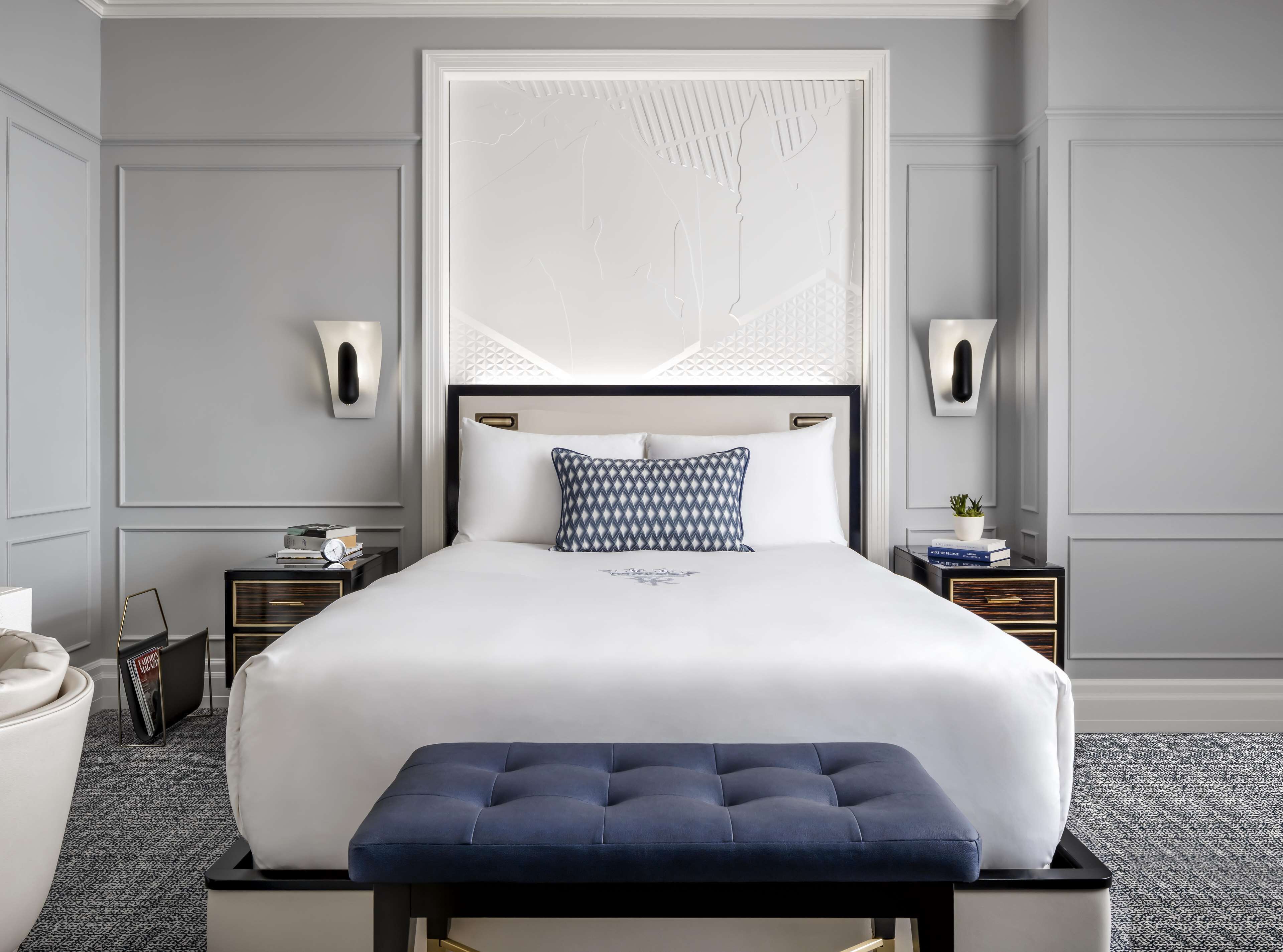 A neatly arranged bedroom with a large bed, a bedside table with a lamp, a small table with a plant, and a bench in front of the bed.