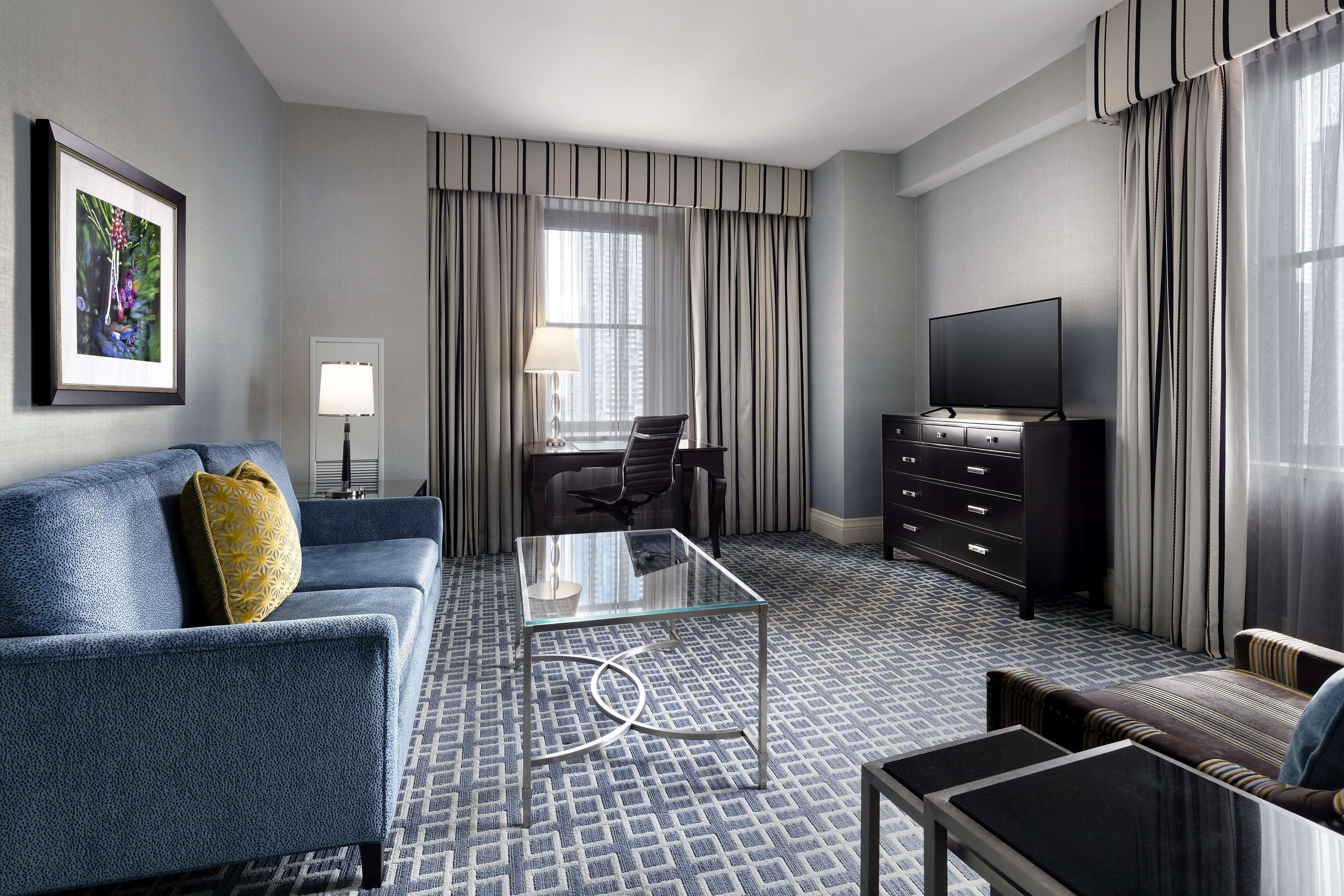 Een hotelkamer met een blauwe bank, een glazen salontafel, een televisie op een dressoir en een kamerbreed tapijt met dessin.