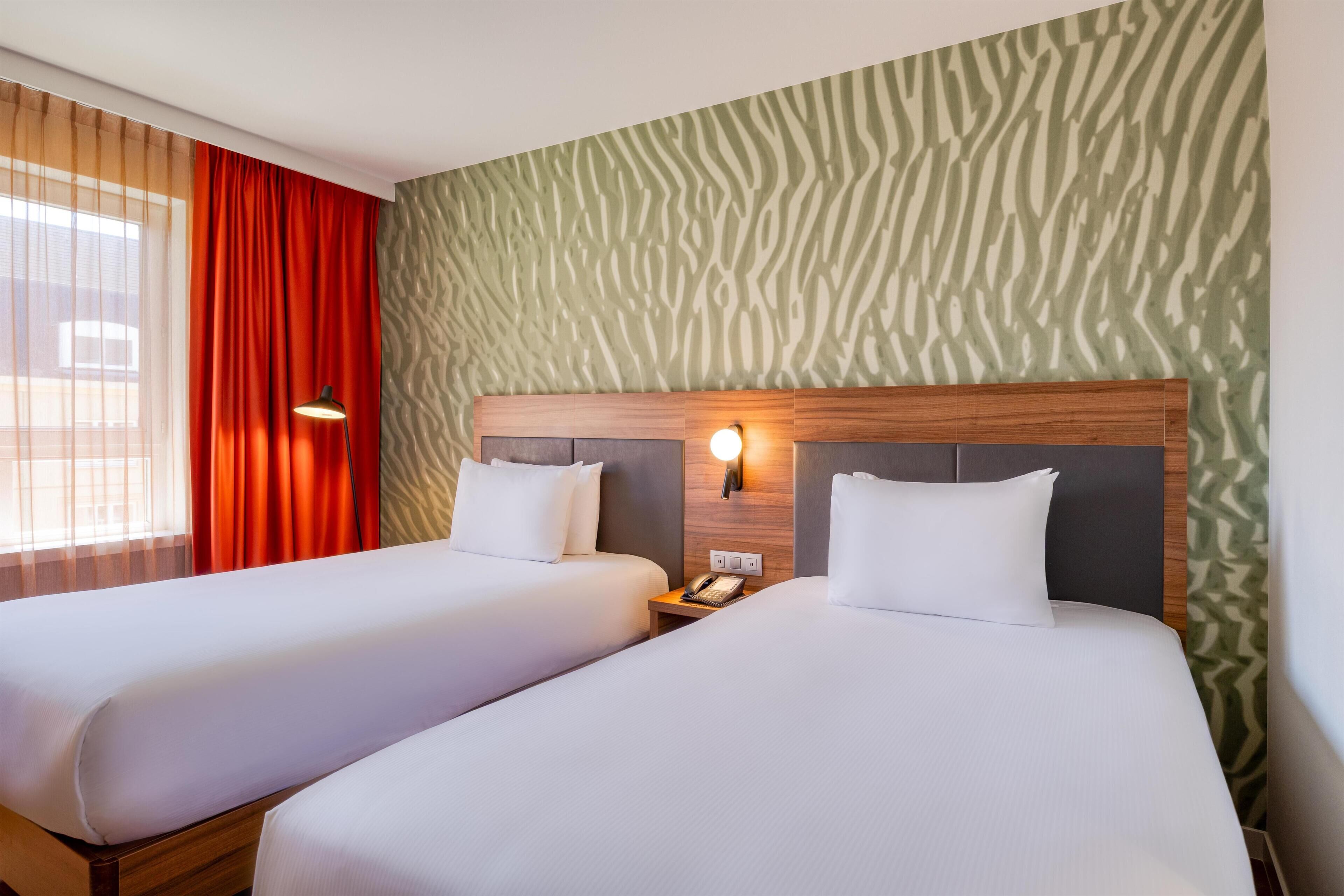 A hotel room with two beds, a patterned wall, and a window with curtains.