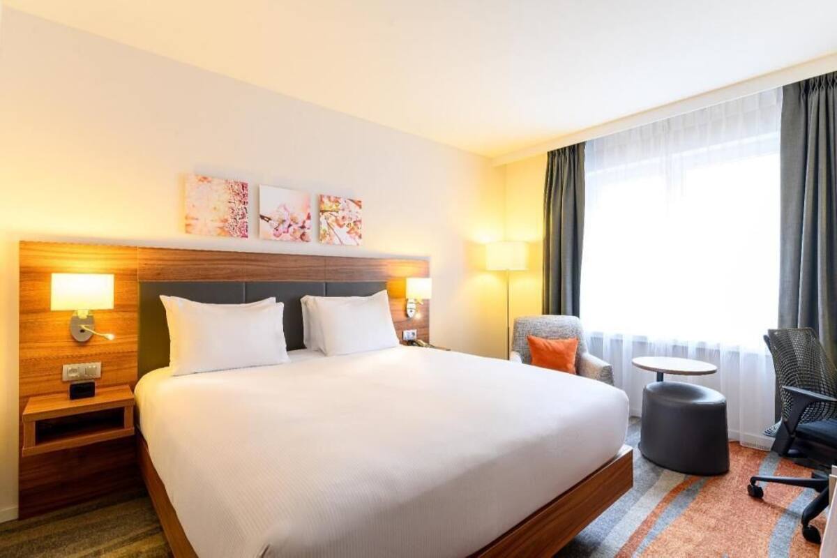 A hotel room with a large bed, bedside lamps, a small table, a chair, and artwork on the wall.