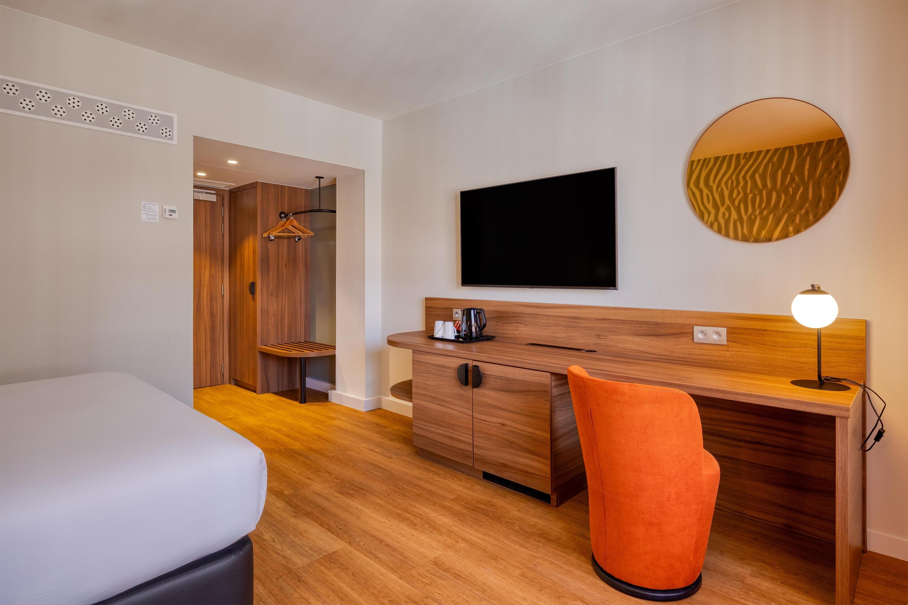 A hotel room with a bed, a desk with a chair, a television, a round mirror, and a wall-mounted light.